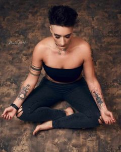Kobieta medytująca w pozycji siedzącej na tle o teksturze kamienia, ubrana w czarny top i dżinsy, z tatuażami na rękach, promuje wydarzenie 'Dzień dla Pań' w Saunie Olimpia