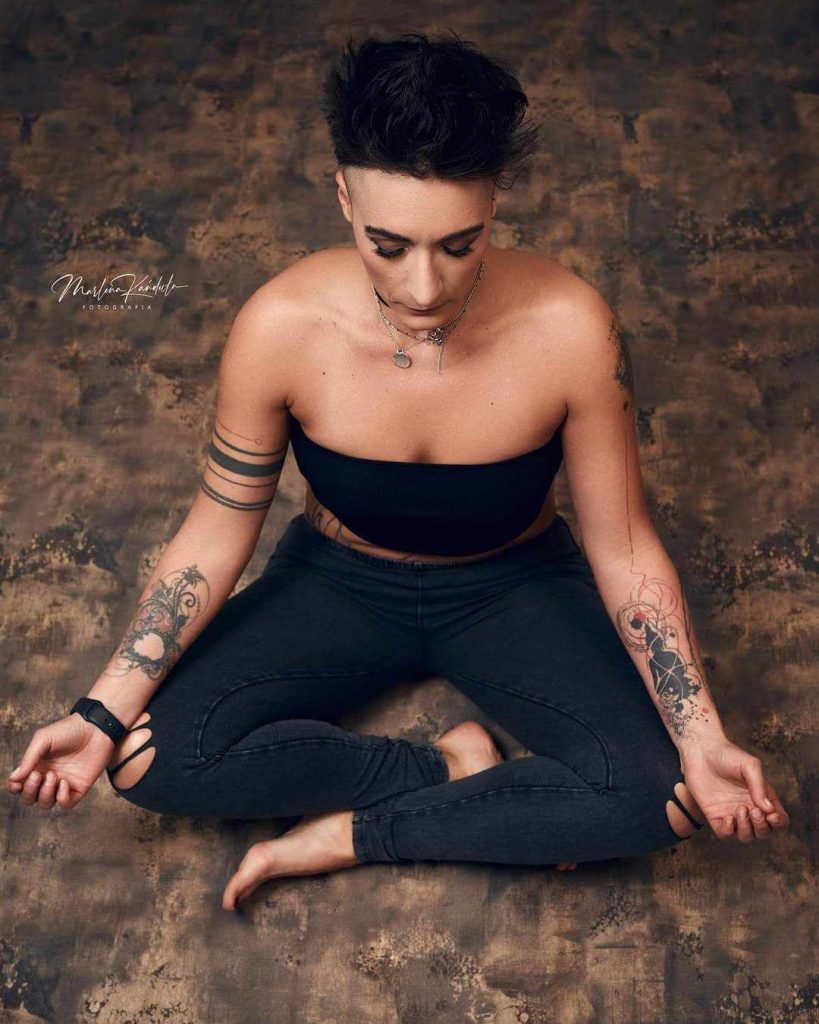 Kobieta medytująca w pozycji siedzącej na tle o teksturze kamienia, ubrana w czarny top i dżinsy, z tatuażami na rękach, promuje wydarzenie 'Dzień dla Pań' w Saunie Olimpia, które odbędzie się 29 lutego 2024 roku.