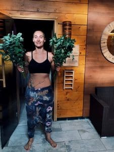 Prowadząca ceremonii saunowej stojąca przed wejściem do sauny z witkami ziołowymi – zapowiedź seansu w Saunie Olimpia.